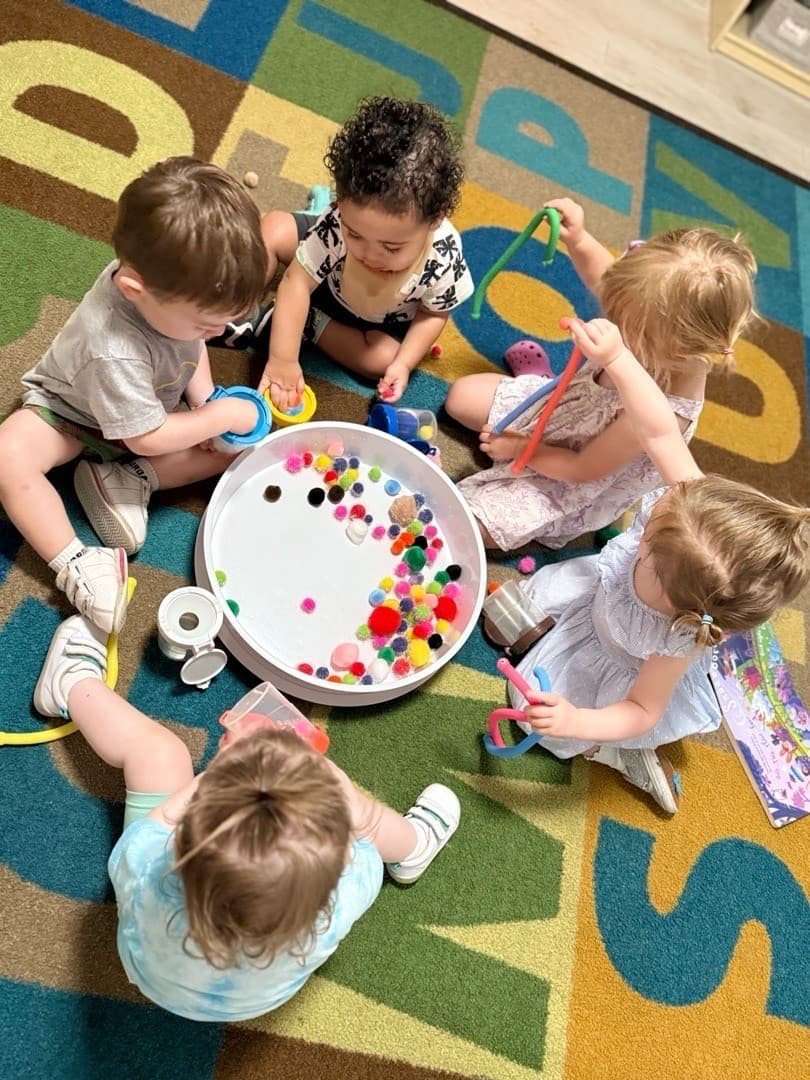 Babies seated together on an alphabet rug exploring colorful pom-poms and sensory tools