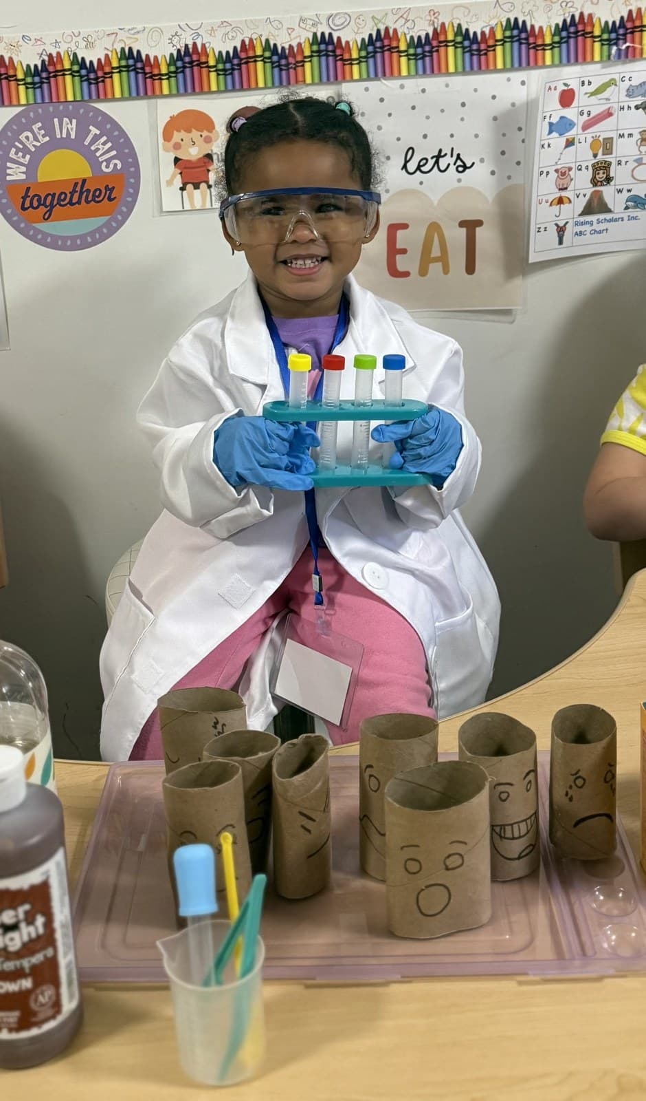 A toddler in a lab coat and goggles holding colorful test tubes during a hands-on science activity