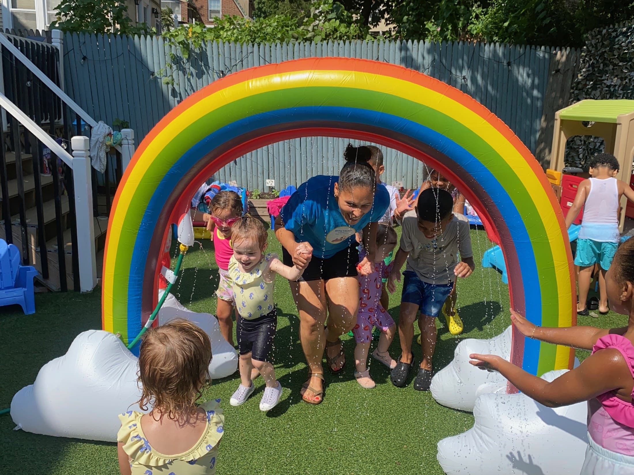 Toddlers and a teacher running through a rainbow sprinkler on the Rising Scholars outdoor play area